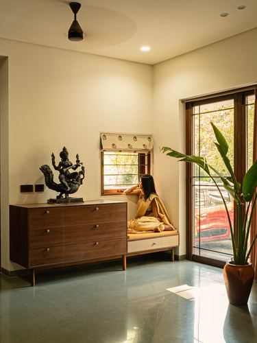 Woman seated on window bench beside wooden dresser with bronze sculpture, sunlit terrazzo floor