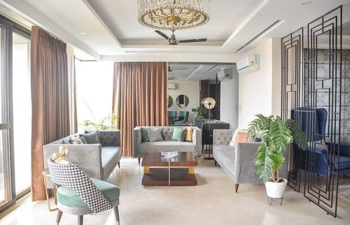Open-plan living room with grey tufted sofas, brass chandelier, and metal jali screen divider
