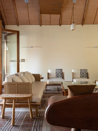 Living room with cane sofa, armchairs, brick flooring, and exposed wooden ceiling under warm light