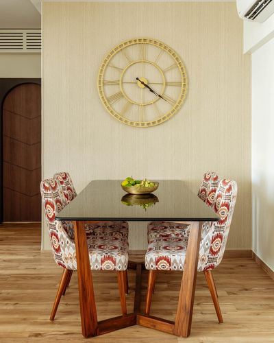 Dining table with glass top and ikat-print chairs beneath a large gold wall clock