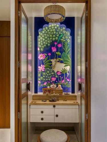Pooja alcove with blue pichwai mural of cow and lotus flowers, brass lamp, white cabinetry.