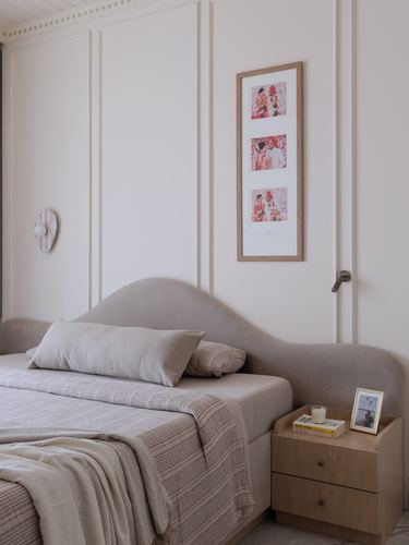 Beige bedroom with curved upholstered headboard, wall paneling, and framed family photos above wooden nightstand.