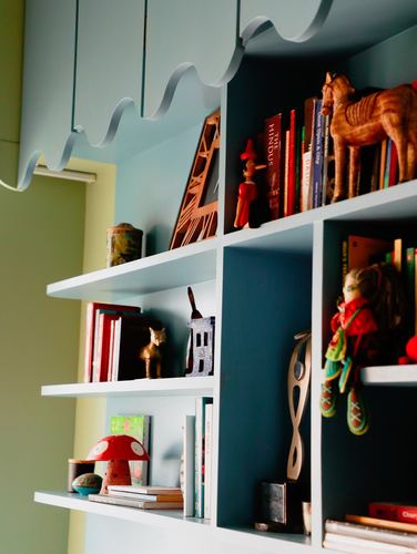 Wall shelving unit with scalloped white trim, teal recesses filled with books, figurines and toys.