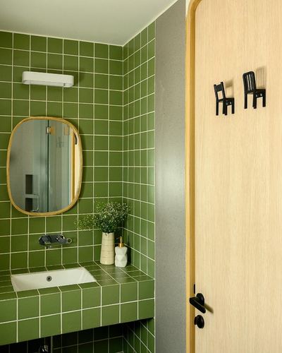 Green tiled bathroom vanity with oval mirror beside a light wood door with chair-shaped hooks