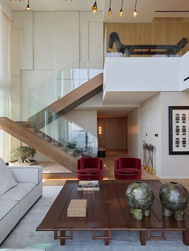 Double-height living room with glass-railed staircase, red velvet chairs, and a dark wood coffee table with ceramic orbs