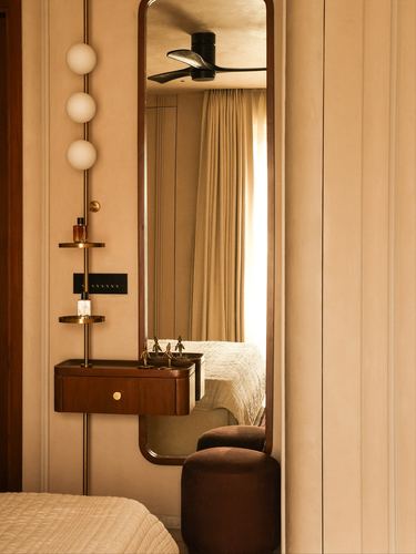 Arched brass mirror above wooden vanity reflects bed and curtained window in warm-toned bedroom