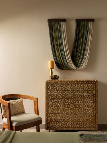 Carved wooden dresser below a woven fiber wall hanging, cane chair beside it