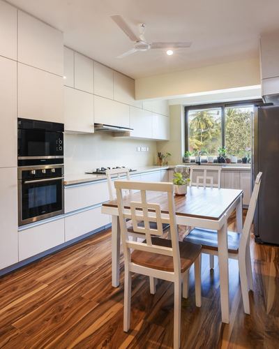 White modern kitchen with wood flooring, dining table, and window view of trees