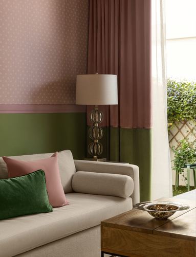 Sofa corner with pink and green pillows, patterned lamp, and pink curtains against a two-tone wall.