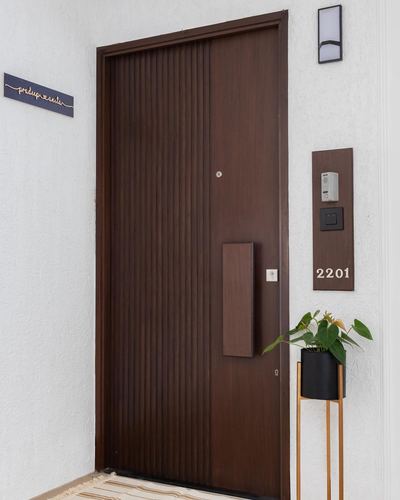 Fluted wooden entry door with brass handle beside numbered plaque and potted plant