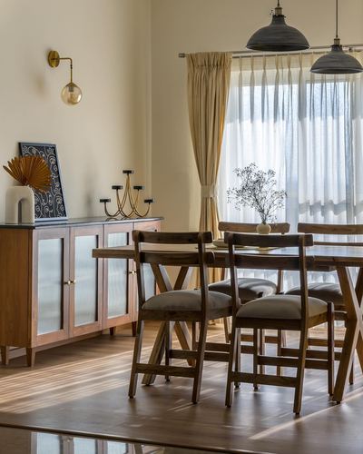 Wooden dining table with cross-back chairs beside sheer-curtained window and fluted glass sideboard