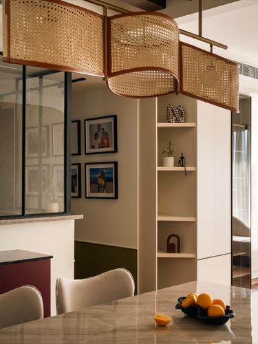 Dining table under a large cane pendant light with a shelving niche and framed photos behind.