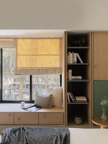 Wooden window seat with cushions beside a bookshelf with green-backed shelves in a Mumbai apartment