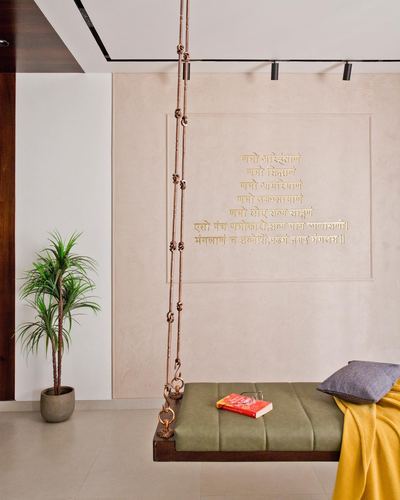 Wooden swing with olive cushion hangs by copper chains before a wall embossed with Sanskrit text