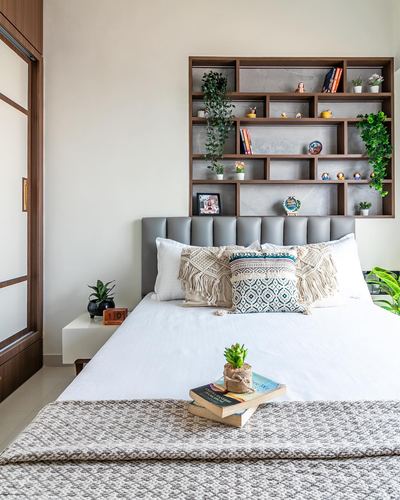 Bedroom with grey quilted headboard, white bedding, patterned throws, and a wooden geometric shelf above filled with plants and decor.