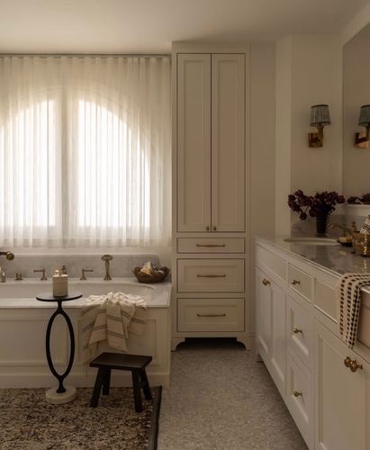 Bathroom with freestanding tub under arched window, cream cabinetry, and brass fixtures