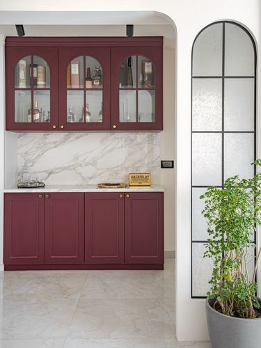 Maroon glass-front bar cabinets with marble backsplash beside an arched fluted glass door and potted plant