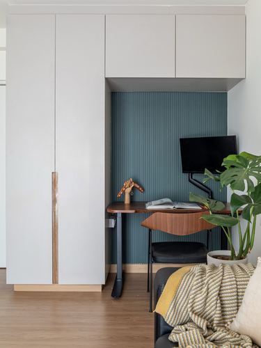 Built-in study nook with ribbed teal panel, wooden desk, monitor arm, and tall wardrobe beside a sofa.