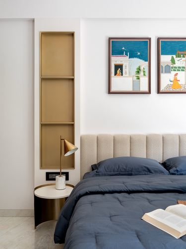 Bedroom corner with brass niche, marble side table, brass lamp, and navy bedding below framed miniature paintings