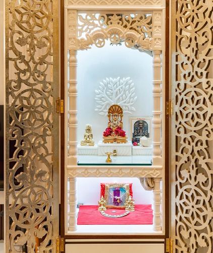 Ornate white pooja mandir with carved jali doors, backlit tree motif, and red cloth diya arrangement below