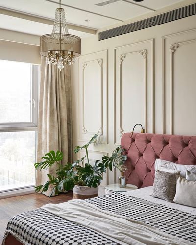 Bedroom corner with pink tufted headboard, panelled wall, chandelier and monstera plants by window