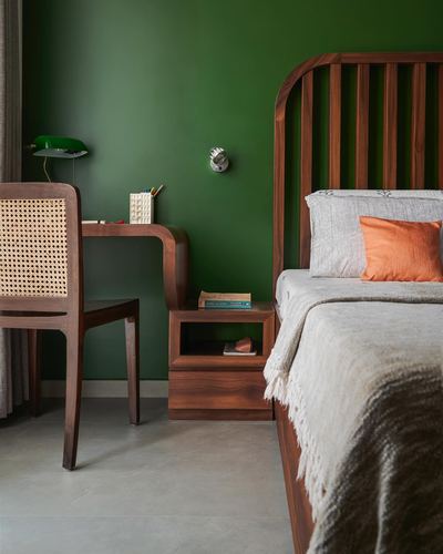 Green bedroom corner with wooden slatted headboard, cane chair, and curved study desk.