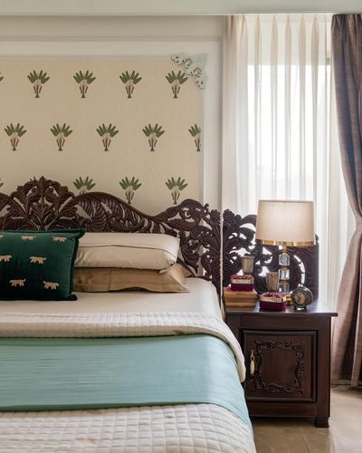 Bed with carved wooden headboard, teal and cream bedding, against botanical wallpaper and window curtains