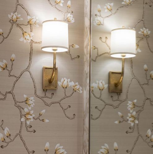 Brass wall sconce with white shade on floral branch wallpaper, reflected in an adjacent mirror panel