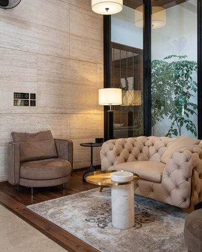Beige living room corner with tufted sofa, round armchair, stone wall, and courtyard glass beyond