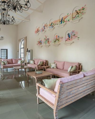 Living room with pink sofas, wooden chairs, and bird murals on tall white wall under chandelier