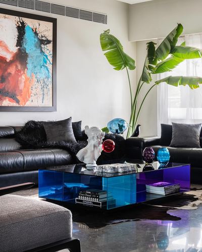 Living room with black leather sofas, blue acrylic coffee table, sculpture bust, banana plant, abstract art