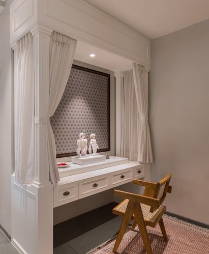 White pooja alcove with curtains, deity idols on marble platform, cane chair beside it