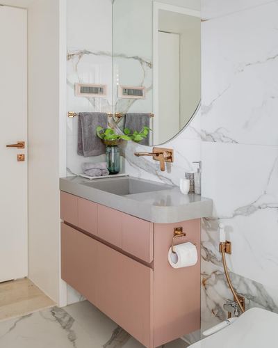 Pink floating vanity with grey stone counter against marble wall, brass fixtures and round mirror