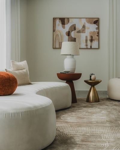 Curved beige sofa with rust pillow beside wood and brass side tables under abstract art