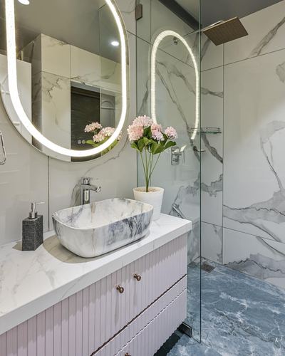Bathroom vanity with marble vessel sink, pink fluted cabinetry, and oval LED mirror beside glass shower