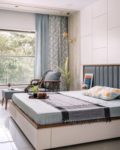 Bedroom with blue tufted headboard, patterned curtains, cane chair, and window view of trees