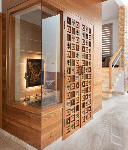 Wooden jali doors beside glass pooja niche with lit deity and floating staircase behind