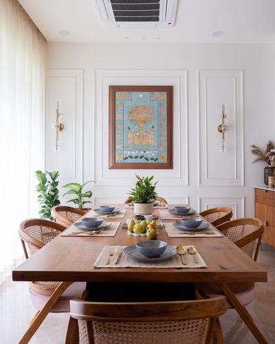 Dining room with wooden cane chairs, table set with blue crockery, and framed pichwai art on wall