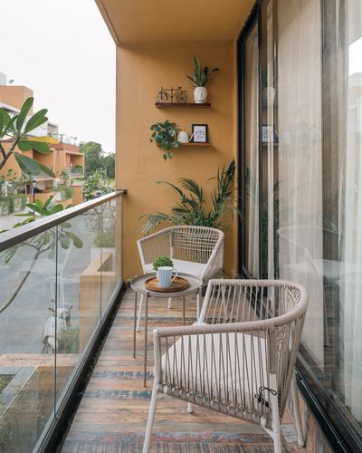 Balcony with mustard wall, woven chairs, wooden floor, plants and glass railing overlooking street