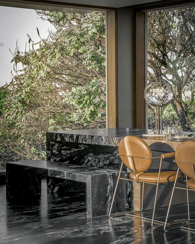 Black marble dining table and bench with tan leather chairs beside floor-to-ceiling corner windows overlooking trees