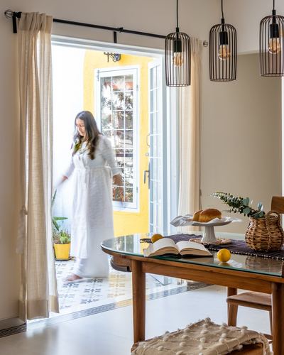 Woman in white robe walking through yellow doorway past a wooden dining table with breakfast setting