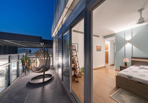 Twilight balcony with hanging egg chair beside glass doors opening into a warmly lit bedroom