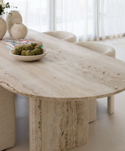 Oval travertine dining table with sculptural legs, styled with vases and a bowl of pinecones