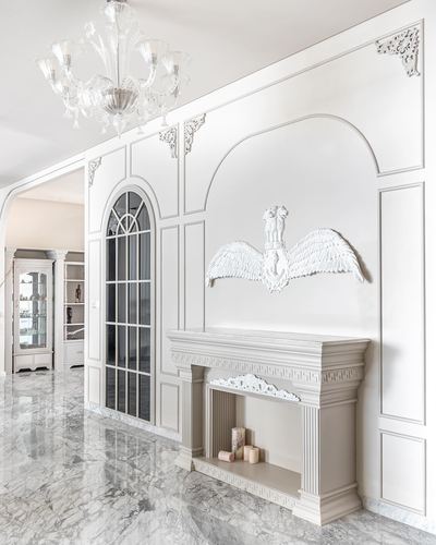 White marble hallway with ornate panelled wall, crystal chandelier, and decorative fireplace mantel with wing sculpture