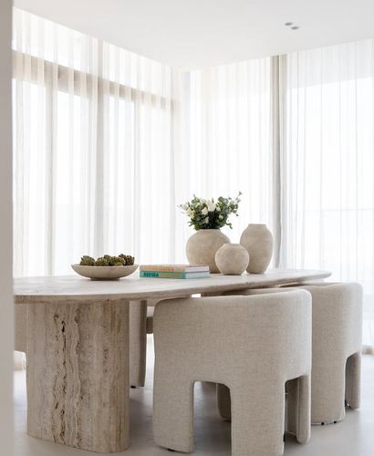 Oval travertine dining table with beige upholstered chairs beside sheer-curtained windows