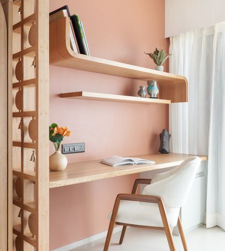 Pink-walled study nook with curved wood shelving, desk, and cane-backed armchair beside sheer curtains