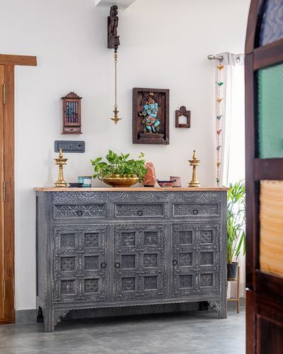 Carved grey wooden sideboard against white wall with hanging brass lamp and framed ganesha carving in Chennai home.