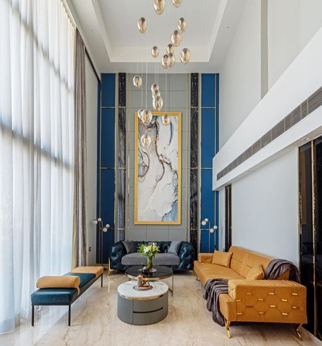 Double-height living room with blue paneled wall, framed artwork, mustard and teal velvet sofas, marble floor