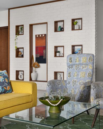 Living room with white brick niche wall, mustard sofa, and printed blue armchairs around a glass table