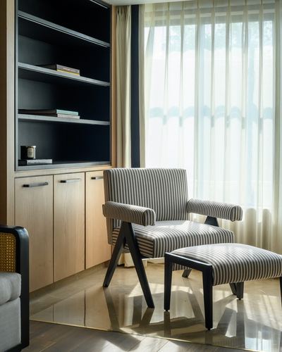 Striped armchair and ottoman by sheer-curtained window beside black and wood shelving unit